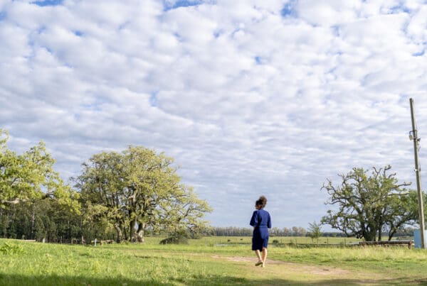 A person in a blue dress walks barefoot across a grassy field under a cloudy sky. Large trees and a pole frame the serene landscape.